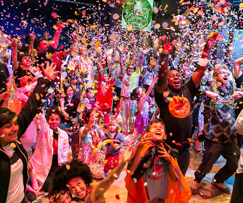 A photo taken a moment after a confetti cannon has gone off at Touretteshero's event Brewing in Battersea in 2018. Children, young people and adults have their hands raised in the air joyfully looking upward and smiling whilst confetti falls all around them. Credit: Kevin Moran